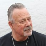 A headshot of a white man with gray hair wearing a black t-shirt, in front of a body of water on an overcast day.