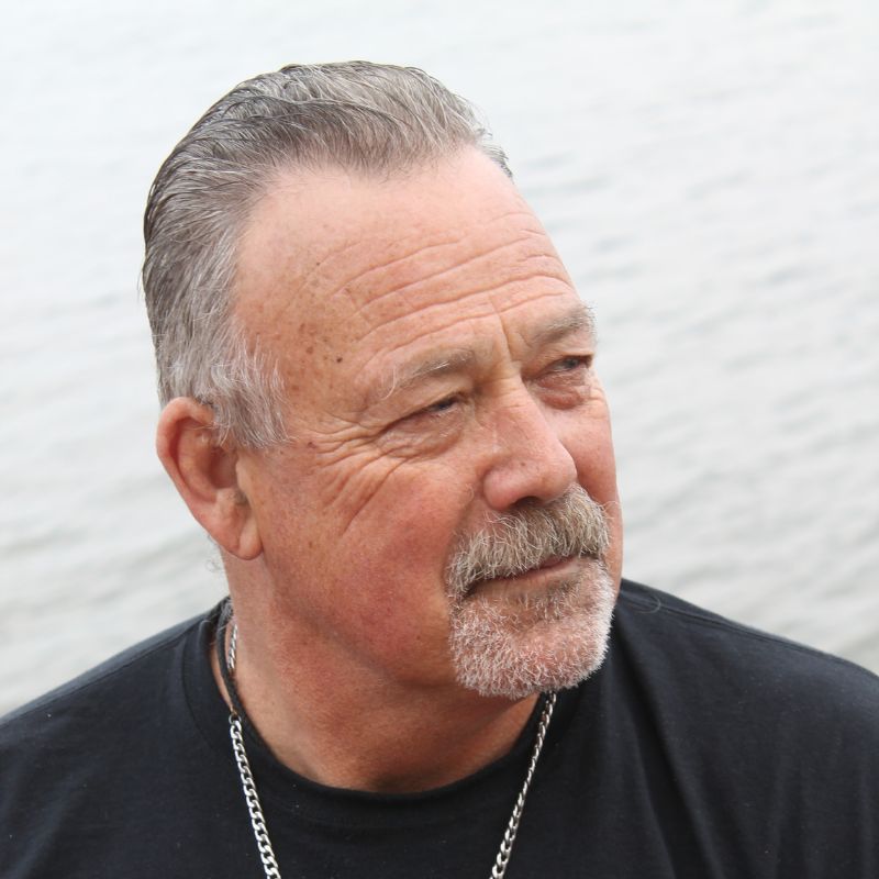 A headshot of a white man with gray hair wearing a black t-shirt, in front of a body of water on an overcast day.