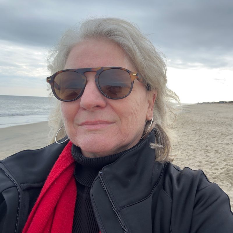 A white woman with gray hair on a beach, on an overcast day. She is wearing a black puffer coat, a red scarf, and sunglasses.