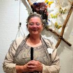 A white woman in front of a sculpture hung on a white wall, made of branches and butterflies.