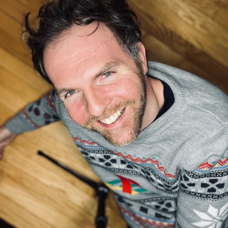 A headshot of a white man wearing a gray sweater, from above, sitting at a wood table in front of a wood wall.