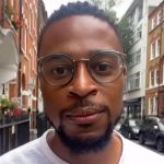 A headshot of a black man wearing glasses and a white shirt, outside in a city.