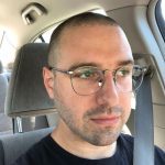 A white man sitting in the car, wearing glasses and a black t-shirt.