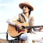 A man with dark hair and a goatee, wearing a hat and brown vest, sitting outside and holding a guitar.
