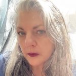 A white woman with long gray hair and dark lipstick, wearing a black shirt.