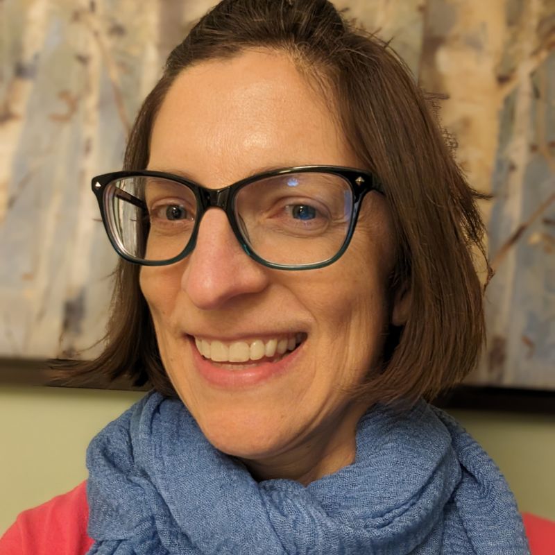 A white woman with a brown bob and glasses, wearing a blue scarf.