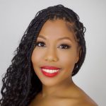 A headshot of a black woman wearing boho braids, a black sleeveless top, and red lipstick, in front of a light gray background.