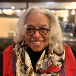 A black woman with long gray hair and a vibrant scarf, wearing glasses and smiling, indoors.