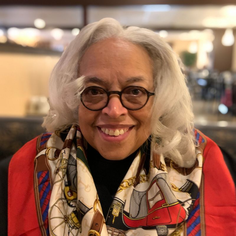 A black woman with long gray hair and a vibrant scarf, wearing glasses and smiling, indoors.